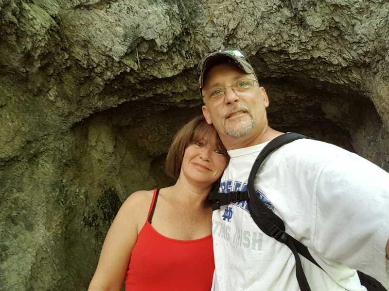 A couple smiling for a selfie in a natural setting, standing close together in front of a rocky cave background. The woman is wearing a red tank top, and the man is dressed in a white shirt with a blue design and a camouflage cap. C&O Canal mountain bike trail.