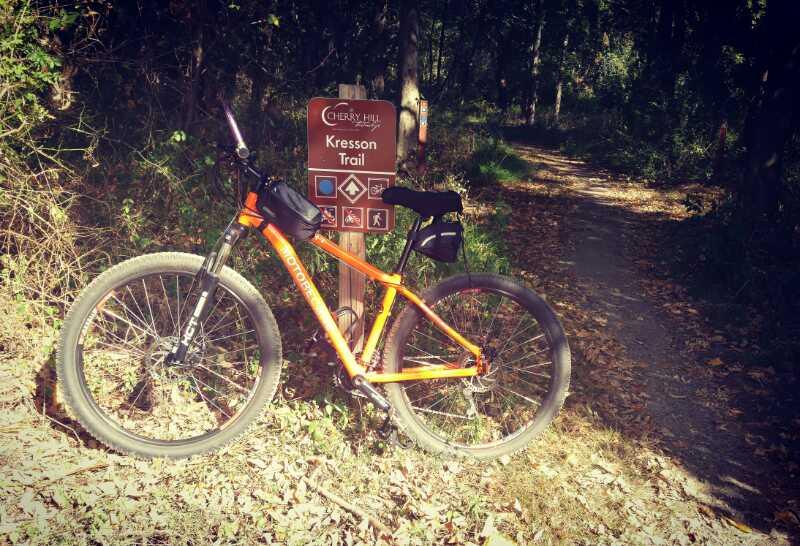 A bright orange mountain bike rests next to a trail sign for Kresson Trail, surrounded by greenery and scattered autumn leaves. The trail leads into a wooded area. Kresson Park mountain bike trail.