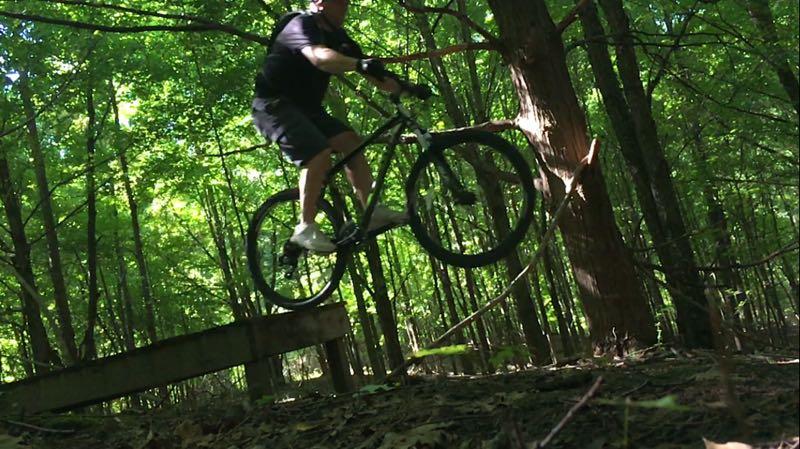 A mountain biker mid-jump on a rugged wooden bridge in a dense, green forest, surrounded by tall trees and sunlight filtering through the leaves. The rider is wearing a black shirt and shorts, with gloves and white shoes, capturing a dynamic moment of action. Anderson Park mountain bike trail.