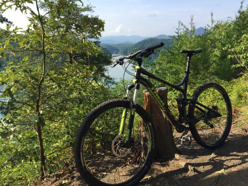 Mountain bike parked near a scenic overlook, surrounded by greenery and trees, with a view of a lake and mountains in the distance under a clear blue sky. Tsali Recreation Area mountain bike trail.