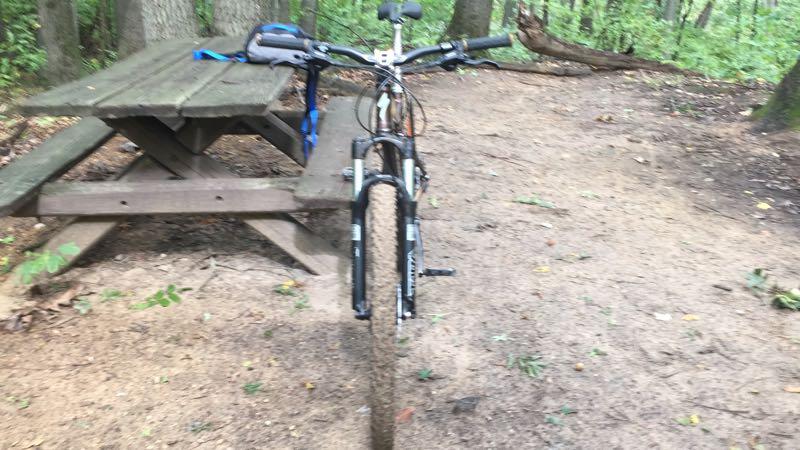 A mountain bike is positioned in the foreground on a dirt path, facing a wooden picnic table in a forested area. The scene features trees in the background, with scattered leaves on the ground, suggesting a natural and serene outdoor environment. Cannonsburg Ski Area mountain bike trail.