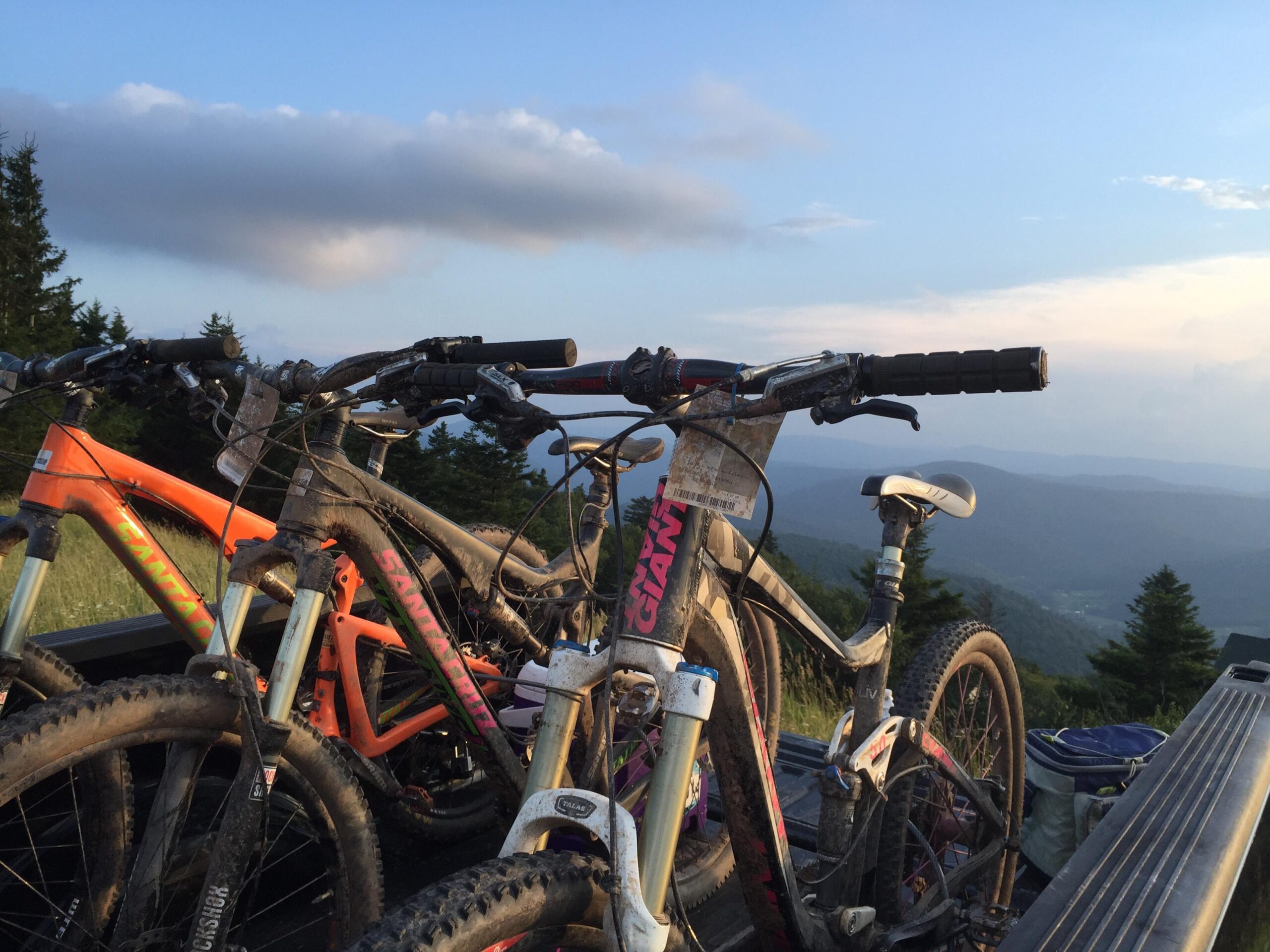Mountain bikes resting on the back of a truck, with a scenic view of rolling hills and trees in the background during sunset. The bikes are muddy, suggesting recent use on rugged trails.