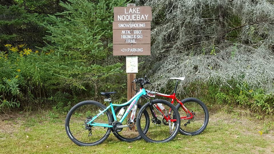 Two mountain bikes, one blue and one red, are parked next to a wooden sign that reads "Lake Noqueabay" with directions for snowshoeing, mountain biking, hiking, and skiing. The sign is surrounded by green foliage and trees. Lake Noquebay mountain bike trail.