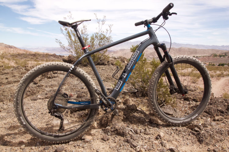 A mountain bike with a sleek gray and blue frame is positioned on rocky terrain, surrounded by sparse vegetation and distant hills. The bike features wide, knobby tires and a modern design, ready for off-road riding. The sky above is partly cloudy, adding to the outdoor adventure ambiance.