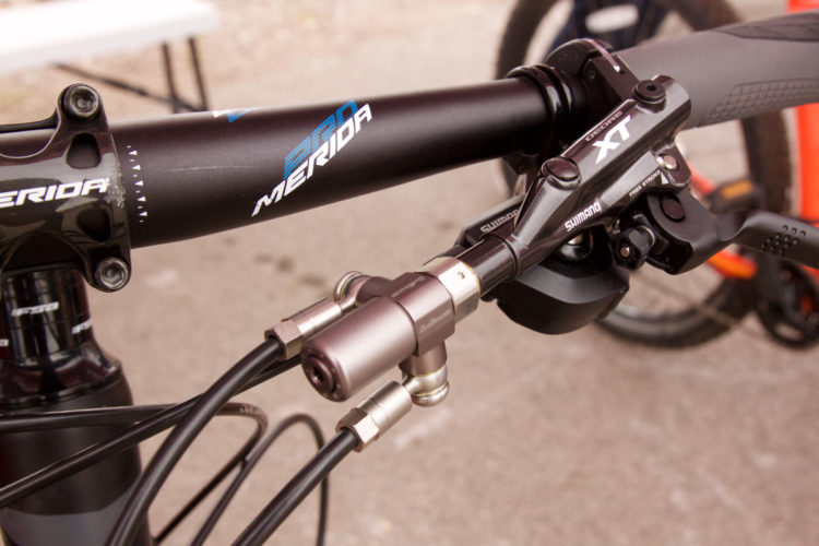 Close-up view of a bicycle handlebar featuring components such as a Shimano XT brake lever, cables, and a Merida branded handlebar. The image highlights the intricate details of the bike's braking system and overall design.
