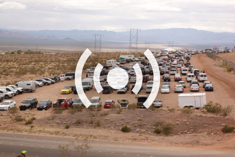 Aerial view of a large parking area filled with numerous vehicles, including cars and vans, situated in a desert landscape. In the background, hills and power lines are visible under a cloudy sky. A sound icon is superimposed in the center of the image.