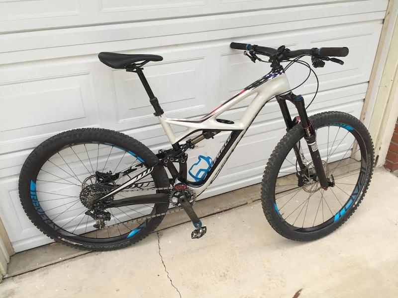 Specialized Enduro Comp 29: A mountain bike with a lightweight silver and black frame, featuring blue accents on the wheels and frame. The bike is parked on a concrete surface in front of a closed white garage door, showcasing its front suspension and gear system.