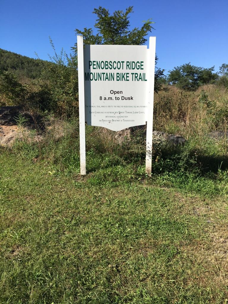 Sign for the Penobscot Ridge Mountain Bike Trail, stating it is open from 8 a.m. to dusk, surrounded by grassy land and trees on a clear day. Nanticoke/wanamie  Penobscot Ridge Mtb Trail mountain bike trail.