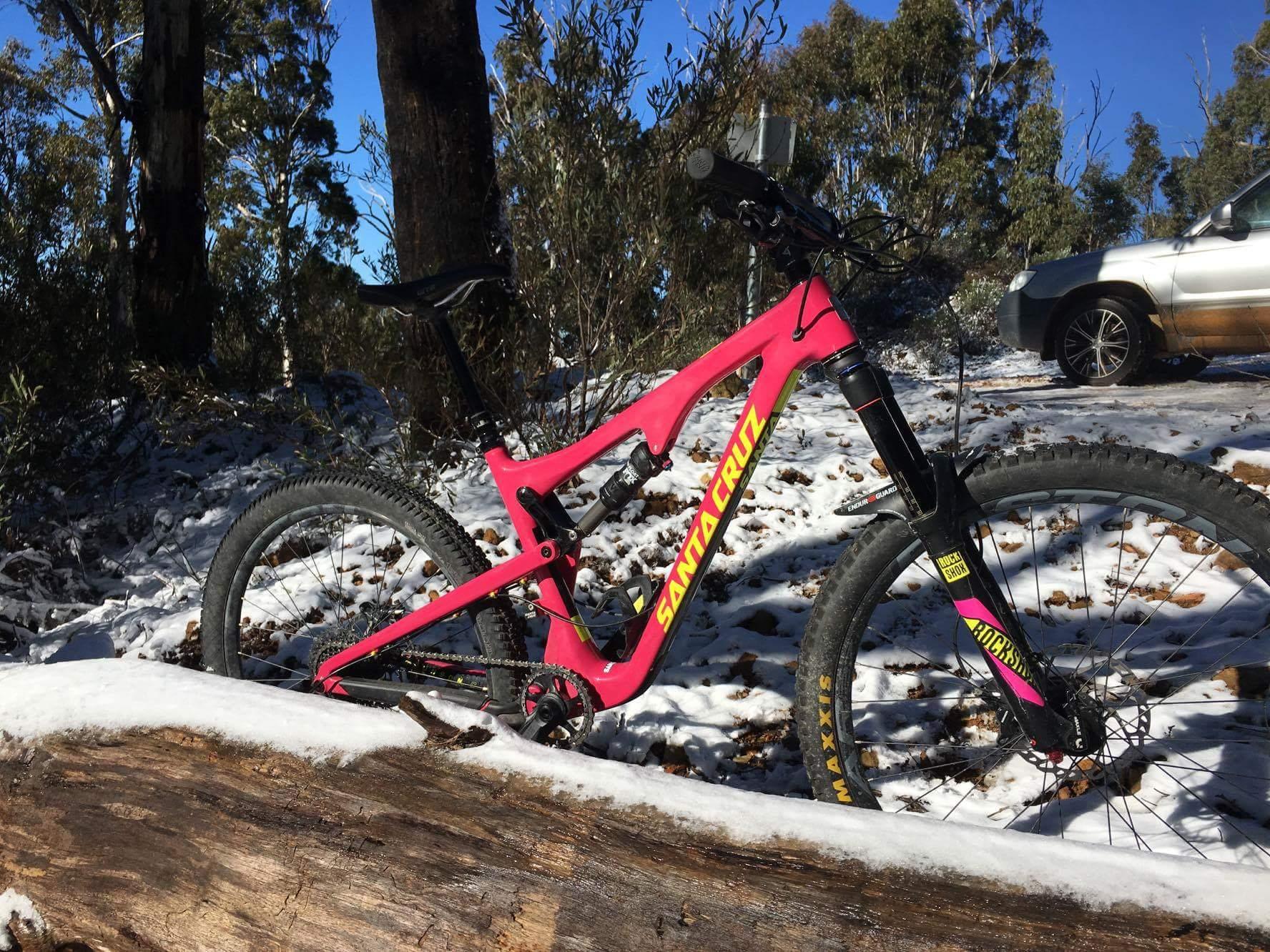 Santa Cruz Bronson 2 CC: A bright pink mountain bike with "Santa Cruz" branding is resting against a log in a snowy landscape. The bike features wide tires and is positioned among trees with green foliage, under a clear blue sky. In the background, a silver car is partially visible.
