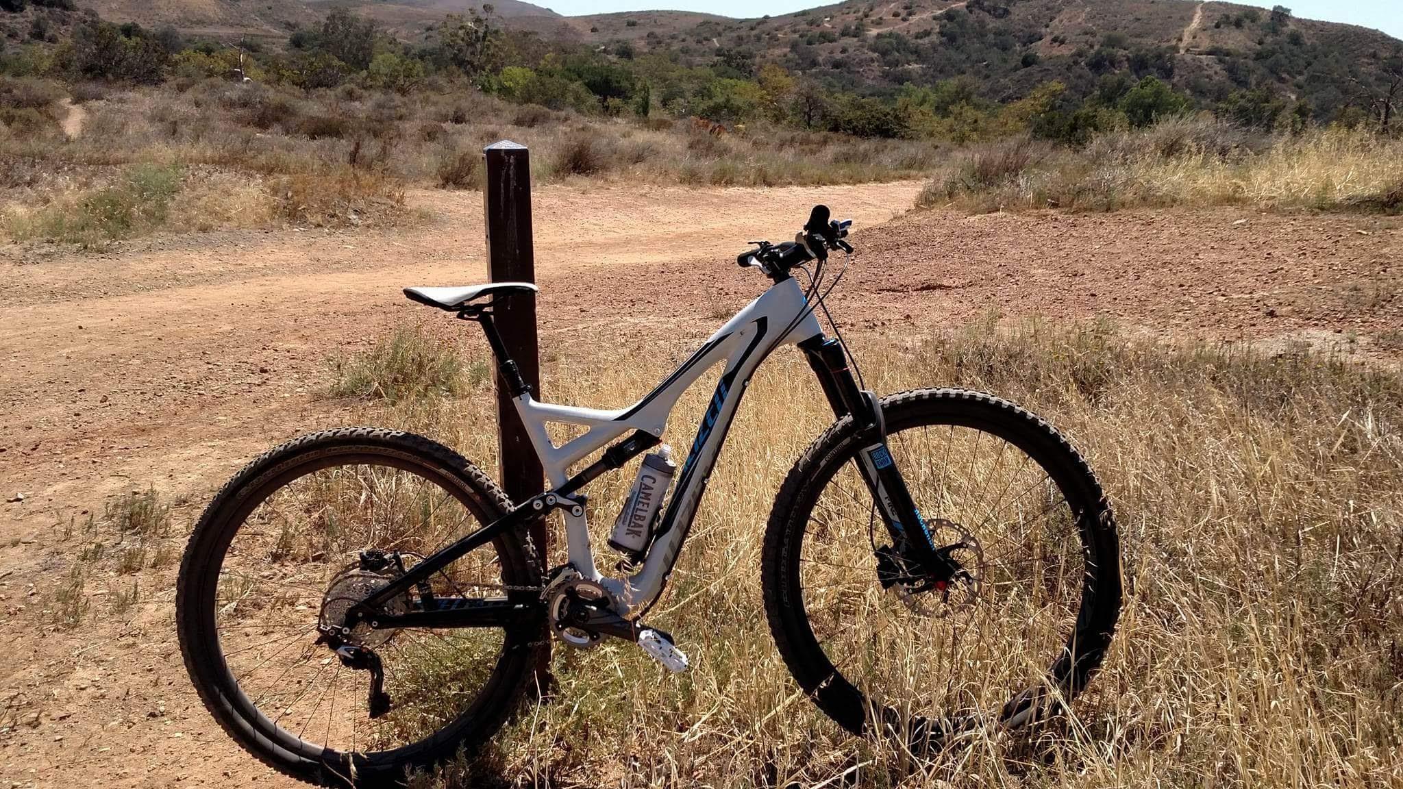 Specialized Stumpjumper FSR Expert: A mountain bike leaning against a wooden post on a dirt trail, surrounded by dry grass and hills in the background under a clear sky.