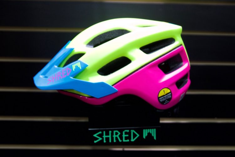 Brightly colored cycling helmet featuring a neon yellow and pink design, with blue accents and the brand name "SHRED" visible on the front. The helmet is mounted on a display stand against a dark background.