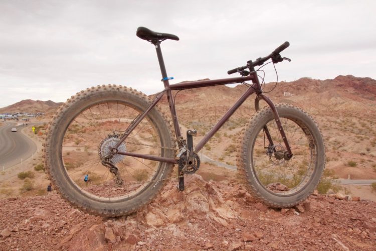 A fat tire bike positioned on a rocky outcrop with a desert landscape in the background. The scene includes rolling hills and a winding road, with a cloudy sky overhead. The bike features large, knobby tires and is designed for off-road riding.