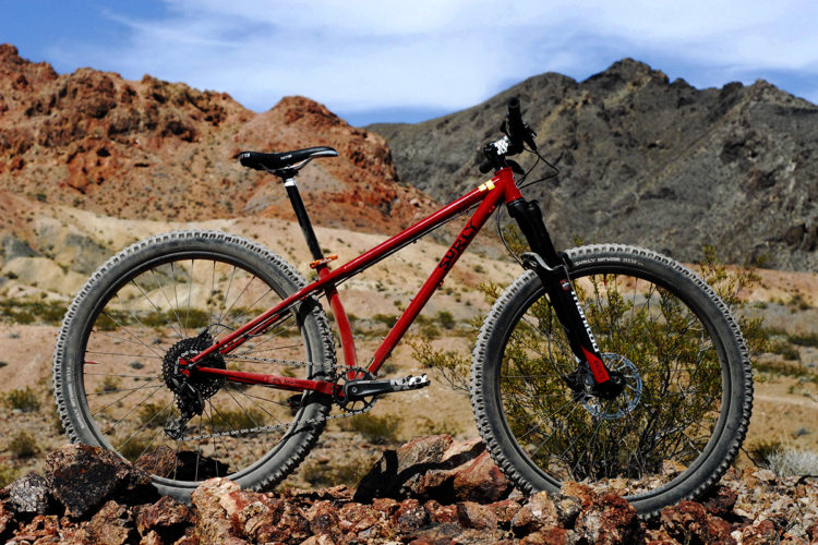 A red mountain bike positioned on rocky terrain, with a backdrop of rugged mountains and a blue sky. The bike features thick tires and a sturdy frame, indicating suitability for off-road trails.