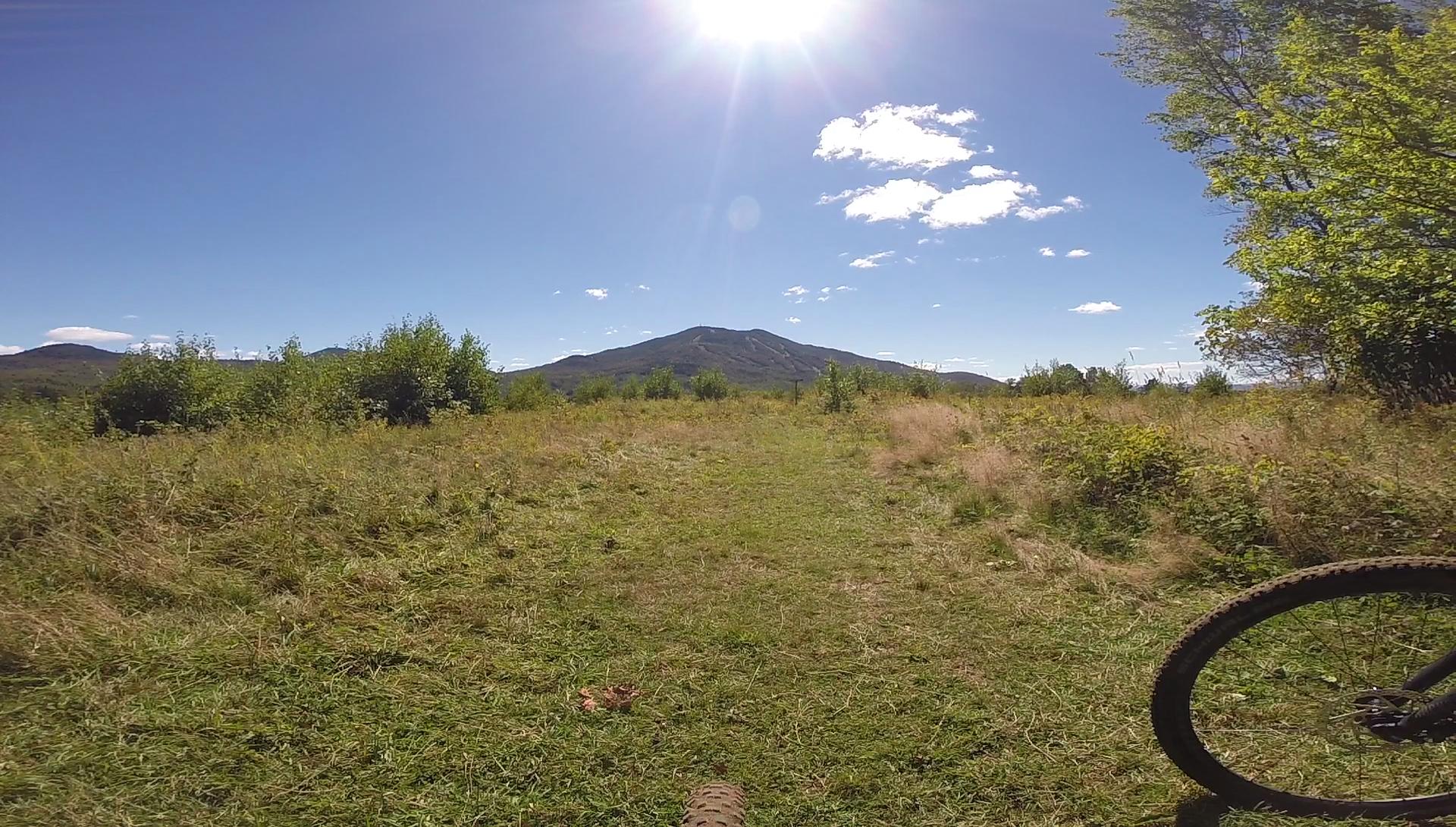 A scenic view of a grassy path leading towards a mountain, under a clear blue sky with the sun shining brightly. The foreground shows part of a bicycle's wheel, hinting at a cycling adventure. In the background, the mountain is framed by patches of green vegetation and distant hills. Kingdom Trails mountain bike trail.