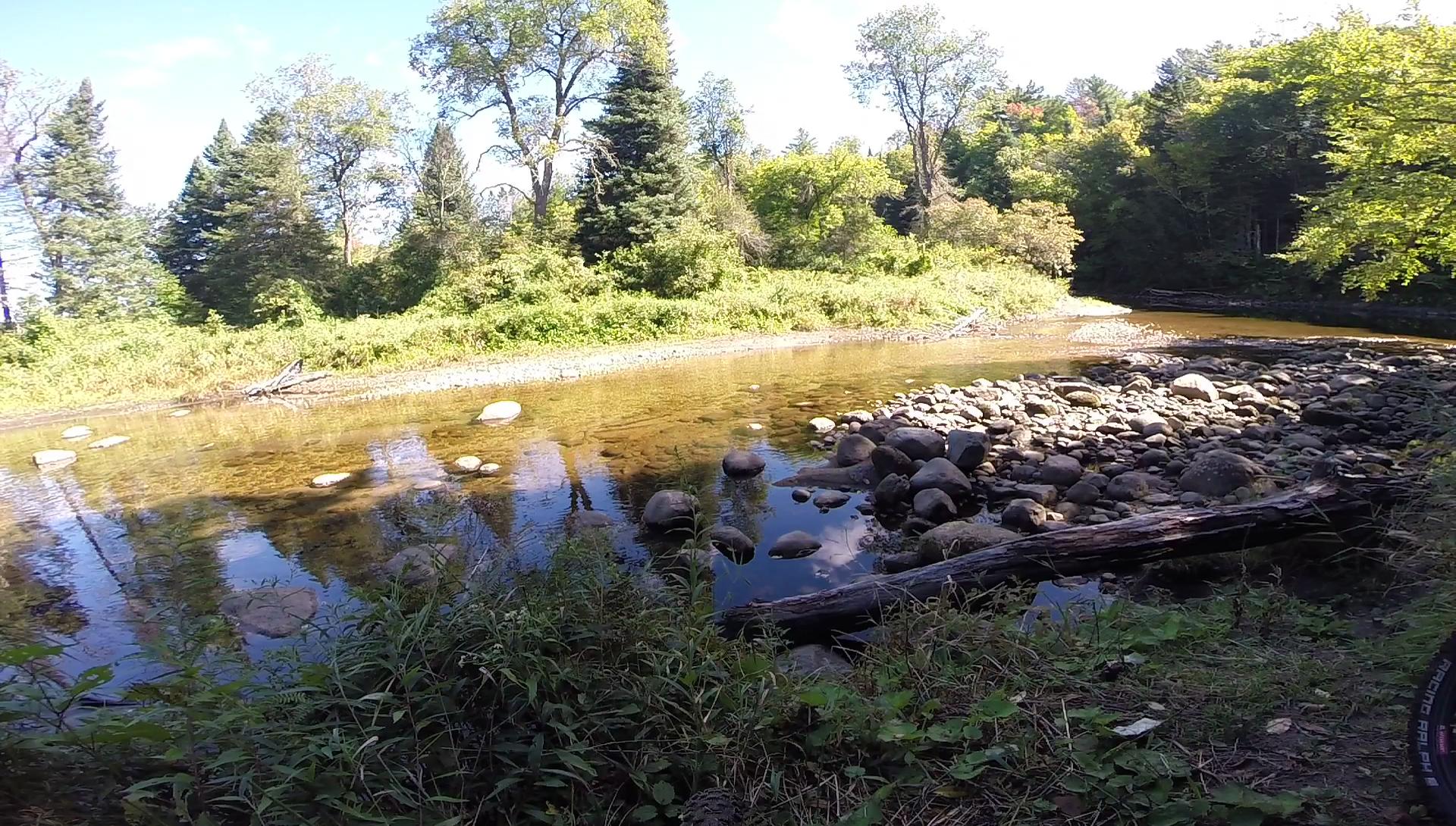 A peaceful river scene with clear water reflecting the blue sky. Large rocks and pebbles line the riverbank, surrounded by lush green vegetation and tall trees. Sunlight filters through the leaves, creating a serene natural environment. Kingdom Trails mountain bike trail.