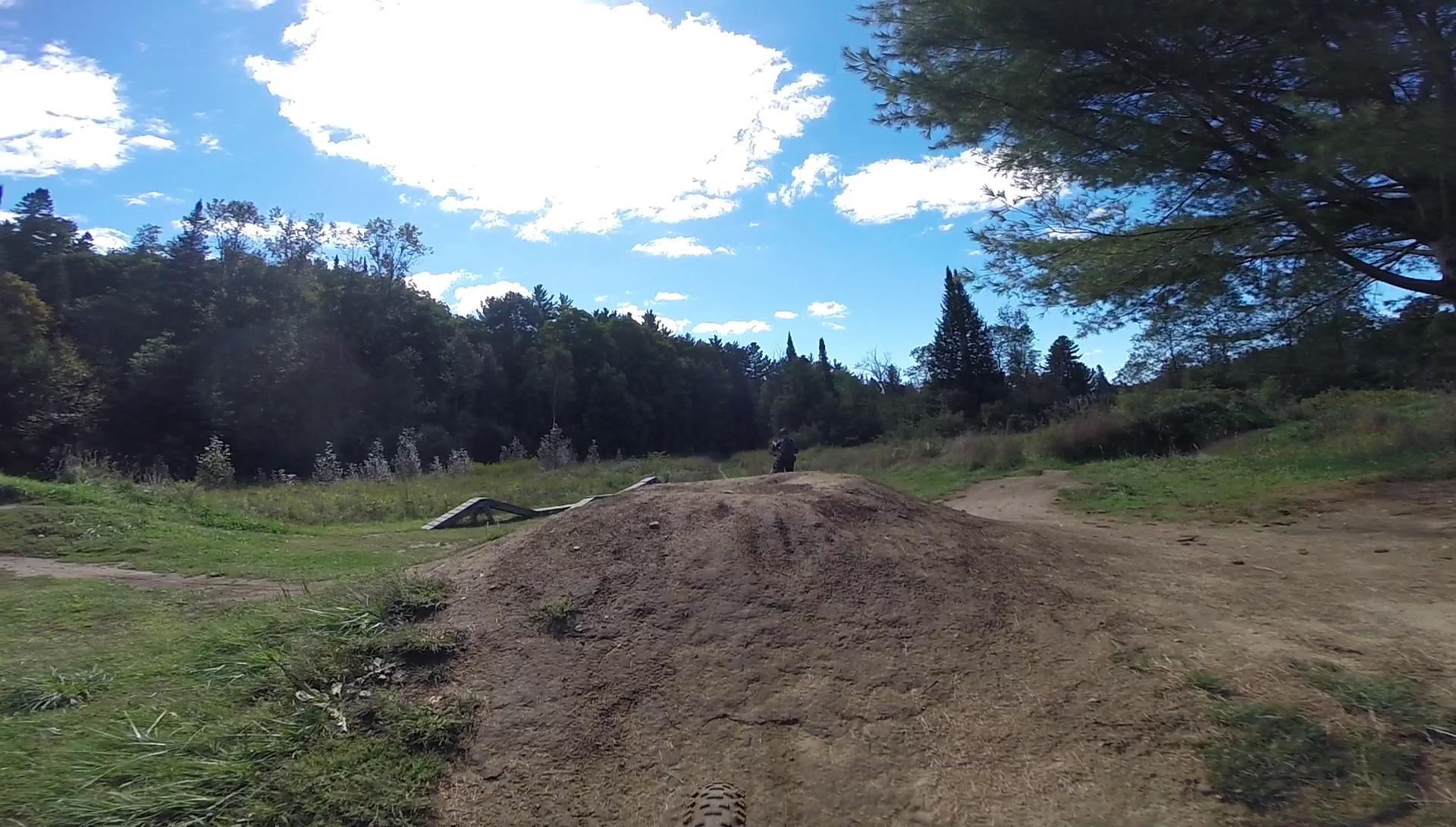 A view of a dirt jump mountain biking trail, featuring a large dirt mound in the foreground, surrounded by green grass and trees. In the background, a rider can be seen approaching the jump, with a blue sky and scattered clouds above. A wooden ramp is visible to the left of the mound. Kingdom Trails mountain bike trail.