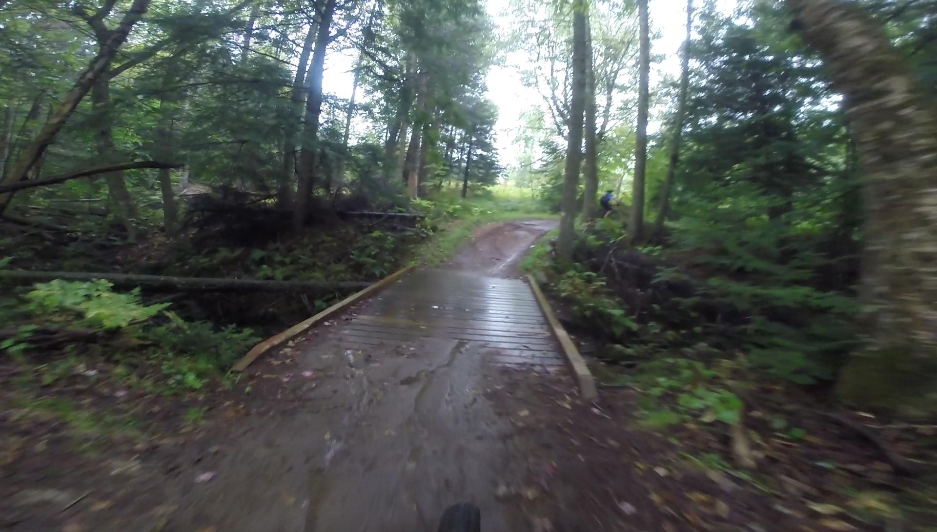 A narrow wooden bridge crossing a muddy path in a lush, green forest. The scene includes tall trees and dense foliage, with a glimpse of a trail leading into the distance. A cyclist can be seen in the background, contributing to the outdoor adventure atmosphere. Kingdom Trails mountain bike trail.