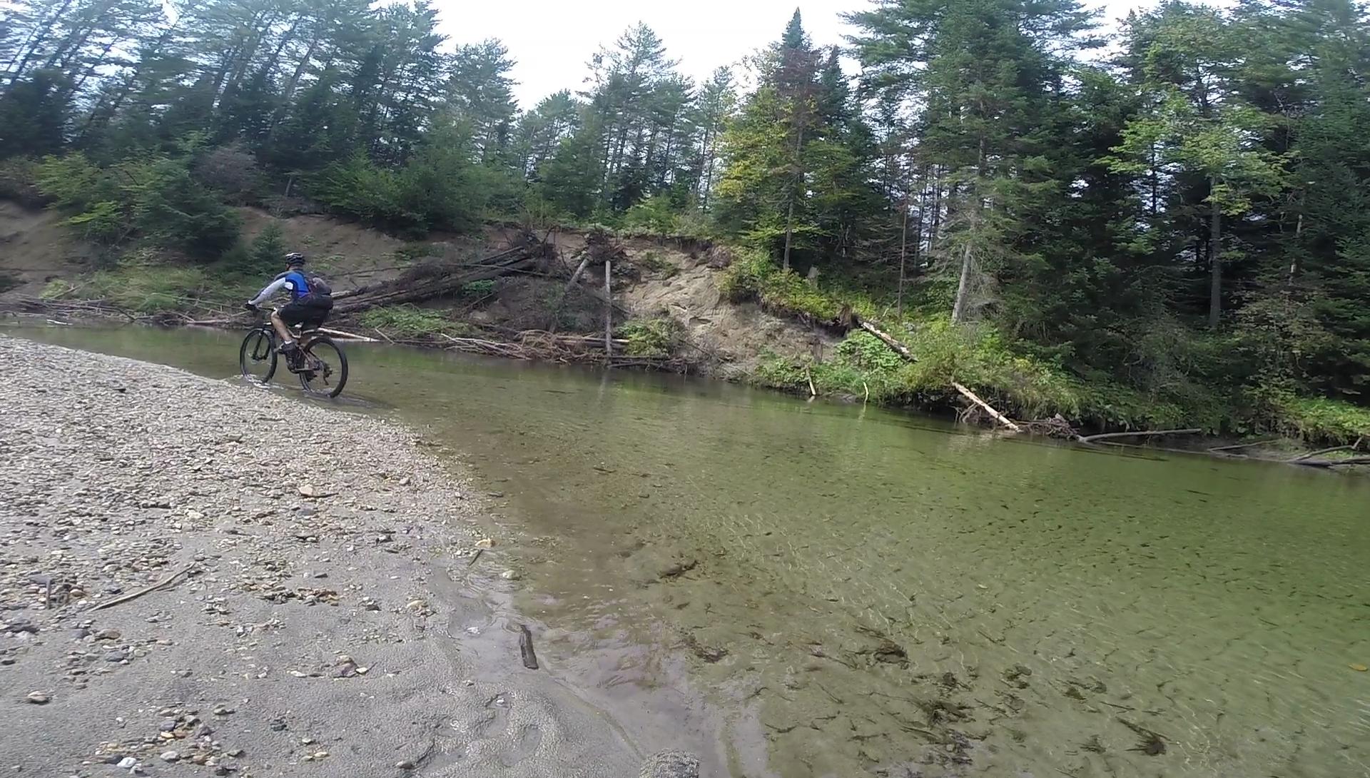 A person rides a mountain bike along a sandy shoreline beside a clear, shallow river, surrounded by dense trees and foliage. The scene captures the beauty of outdoor adventure in a natural setting. Kingdom Trails mountain bike trail.