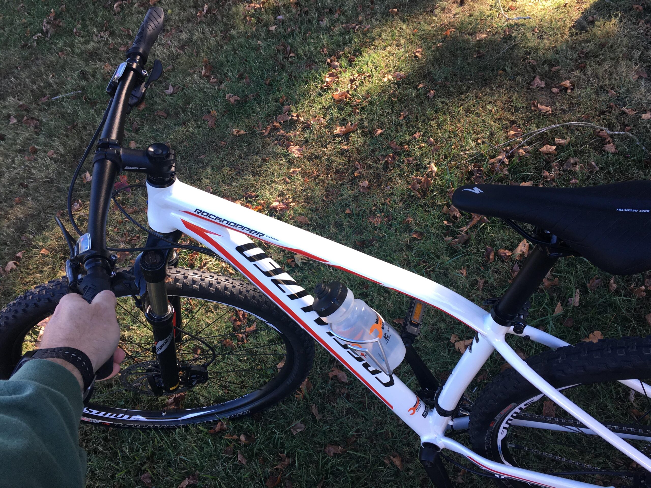 Specialized Rockhopper Comp 29: A close-up image of a mountain bike, showing the handlebars, front wheel, and part of the bike frame. A hand is gripping the handlebars, and a water bottle is mounted on the frame. The bike is positioned on grass with scattered leaves.