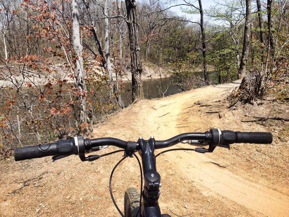 View from the handlebars of a mountain bike on a dirt path beside a river, surrounded by trees with early spring foliage. The trail curves slightly, leading toward a scenic natural landscape. Alum Creek Phase II mountain bike trail.