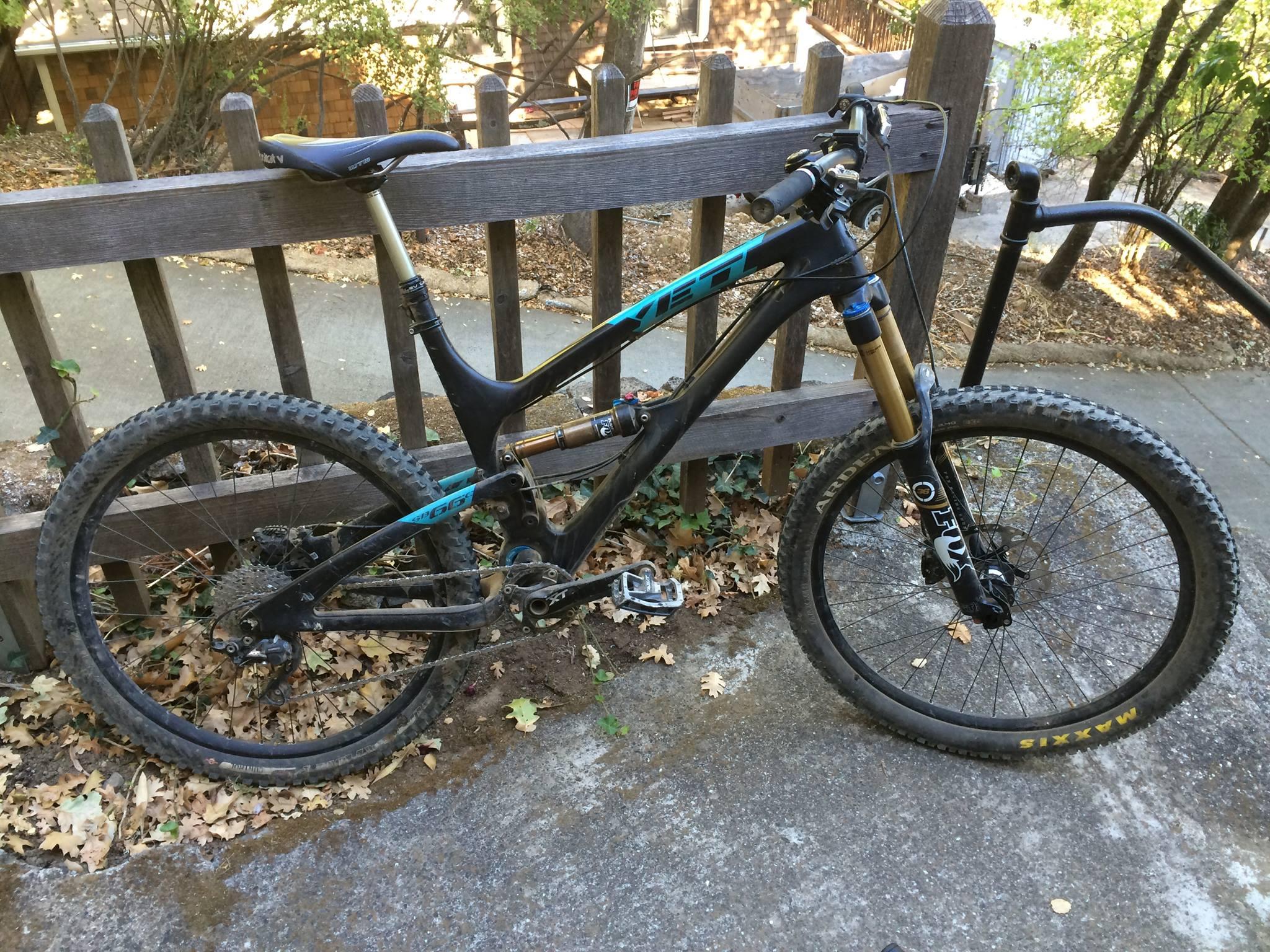 Yeti SB66c: A black mountain bike with blue accents leans against a wooden fence. The bike features wide tires with visible tread patterns, a suspension fork, and a dropper seat post. Fallen leaves cover the ground around the bike, indicating an outdoor setting. The background includes patches of greenery and a blurred building.
