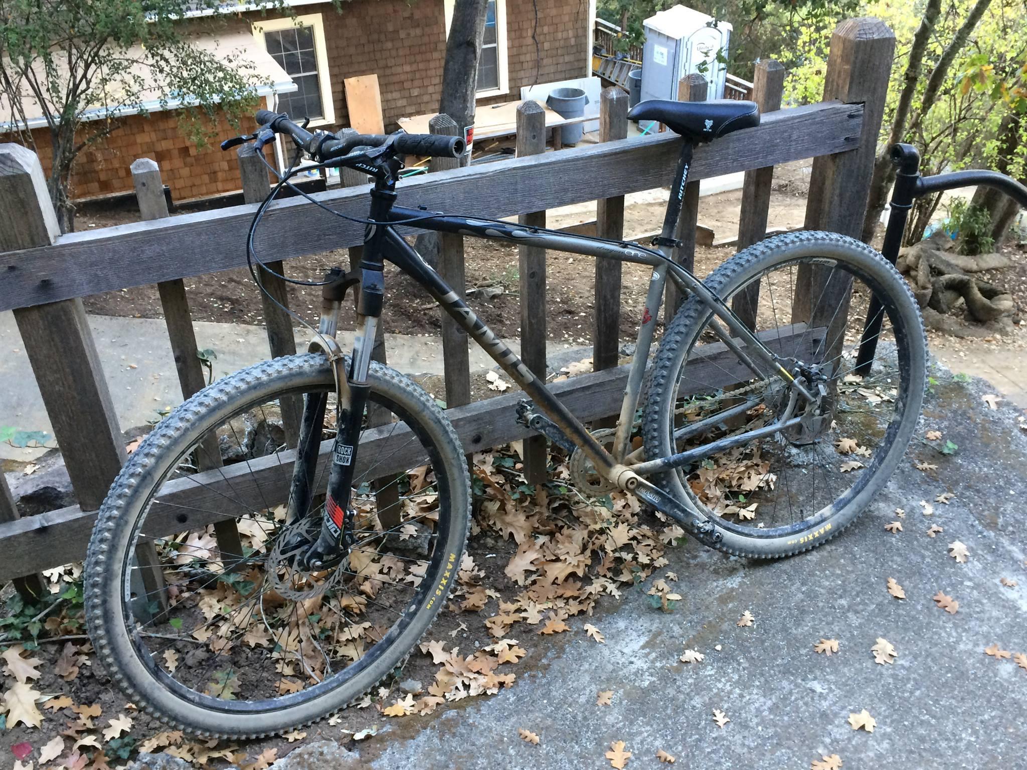 Jamis Dragon: A mountain bike with a black frame leaning against a wooden fence, surrounded by fallen leaves. The bike has noticeable dirt on the tires and frame, indicating recent use on a rugged terrain. In the background, a partially visible wooden building and outdoor structures can be seen.