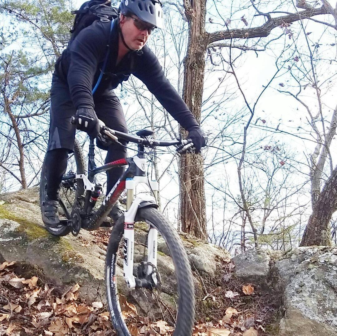 Trek Remedy 9.8 27.5/650b: A cyclist in a black helmet and gear navigates over rough terrain on a mountain bike, surrounded by trees and rocky surfaces. The ground is covered with fallen leaves.