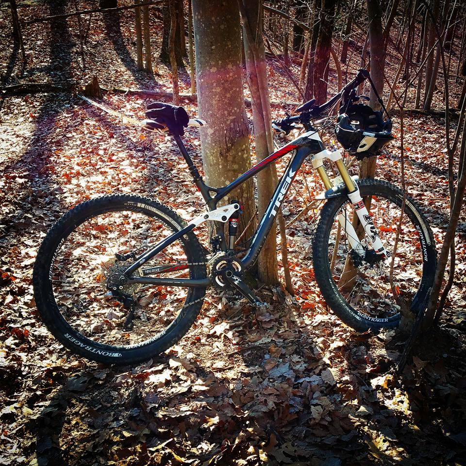 Trek Remedy 9.8 27.5/650b: Mountain bike leaning against a tree in a wooded area, with fallen leaves scattered on the ground. The sunlight creates a warm glow, highlighting the bike's sleek design in black and silver.