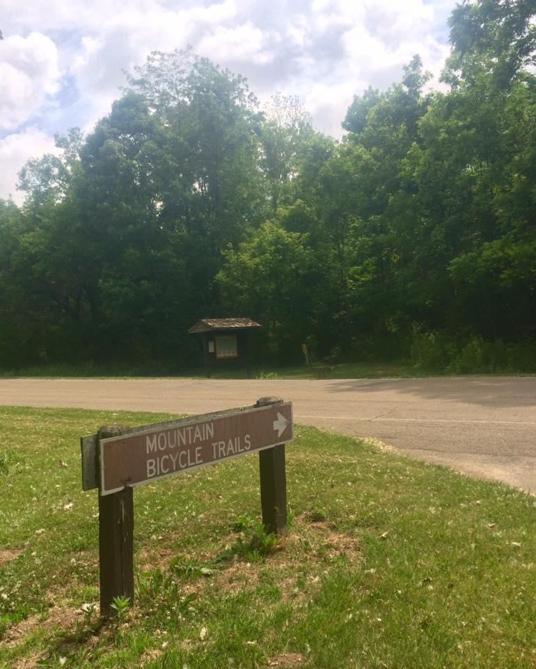 Sign indicating the direction to mountain bicycle trails, surrounded by green trees and a grassy area, under a partly cloudy sky. In the background, there is a wooden information kiosk. Hueston Woods State Park mountain bike trail.