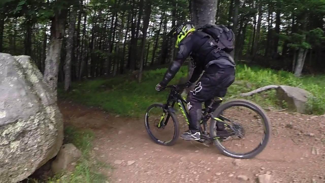 A mountain biker in protective gear rides along a dirt trail in a wooded area, maneuvering around a large rock. The scene features greenery and trees in the background, highlighting the natural setting of the trail. Snowshoe Bike Park mountain bike trail.
