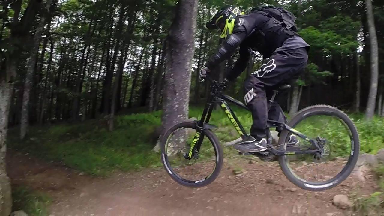 A mountain biker in protective gear jumps off a dirt ramp in a forested area, surrounded by trees and greenery. The biker is wearing a black outfit with accents of yellow, showcasing an adventurous moment in downhill biking. Snowshoe Bike Park mountain bike trail.