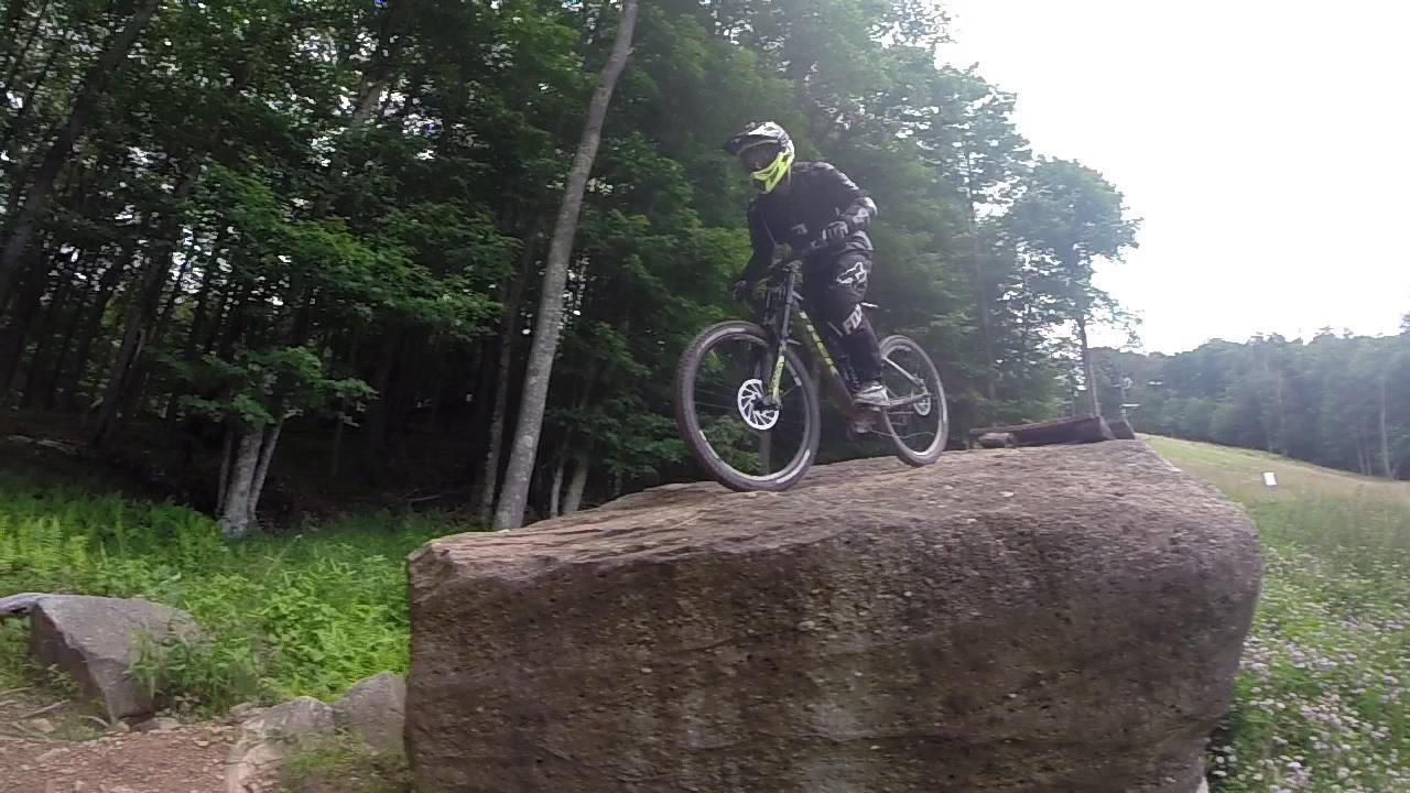 A mountain biker wearing a black outfit and a helmet with a bright yellow visor jumps off a large rock formation in a wooded area, surrounded by trees and greenery. Snowshoe Bike Park mountain bike trail.