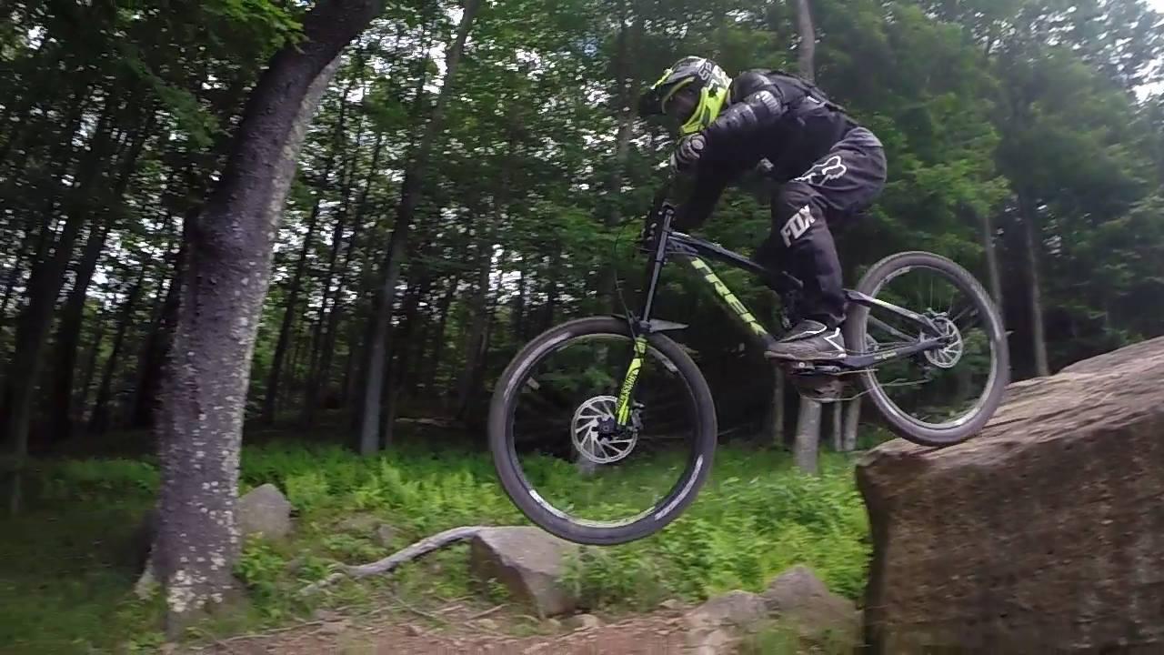A mountain biker in protective gear jumps off a large rock, capturing mid-air action in a forested setting. The surrounding foliage consists of green trees and grass, creating a natural backdrop for the thrilling outdoor sport. Snowshoe Bike Park mountain bike trail.