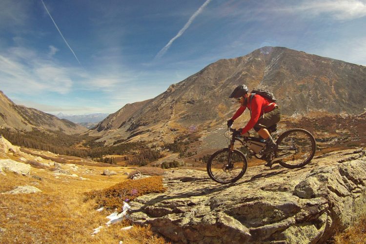 A mountain biker in a red long-sleeve shirt rides over rocky terrain, surrounded by a picturesque landscape of mountains and valleys under a clear blue sky. The scene captures the thrill of outdoor adventure and the natural beauty of the wilderness.
