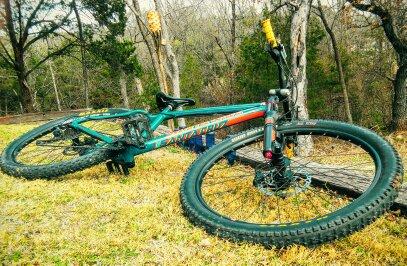 Santa Cruz Chameleon: A teal and orange mountain bike lying on green grass with trees in the background. The bike is positioned on its side, displaying its large tires and frame.