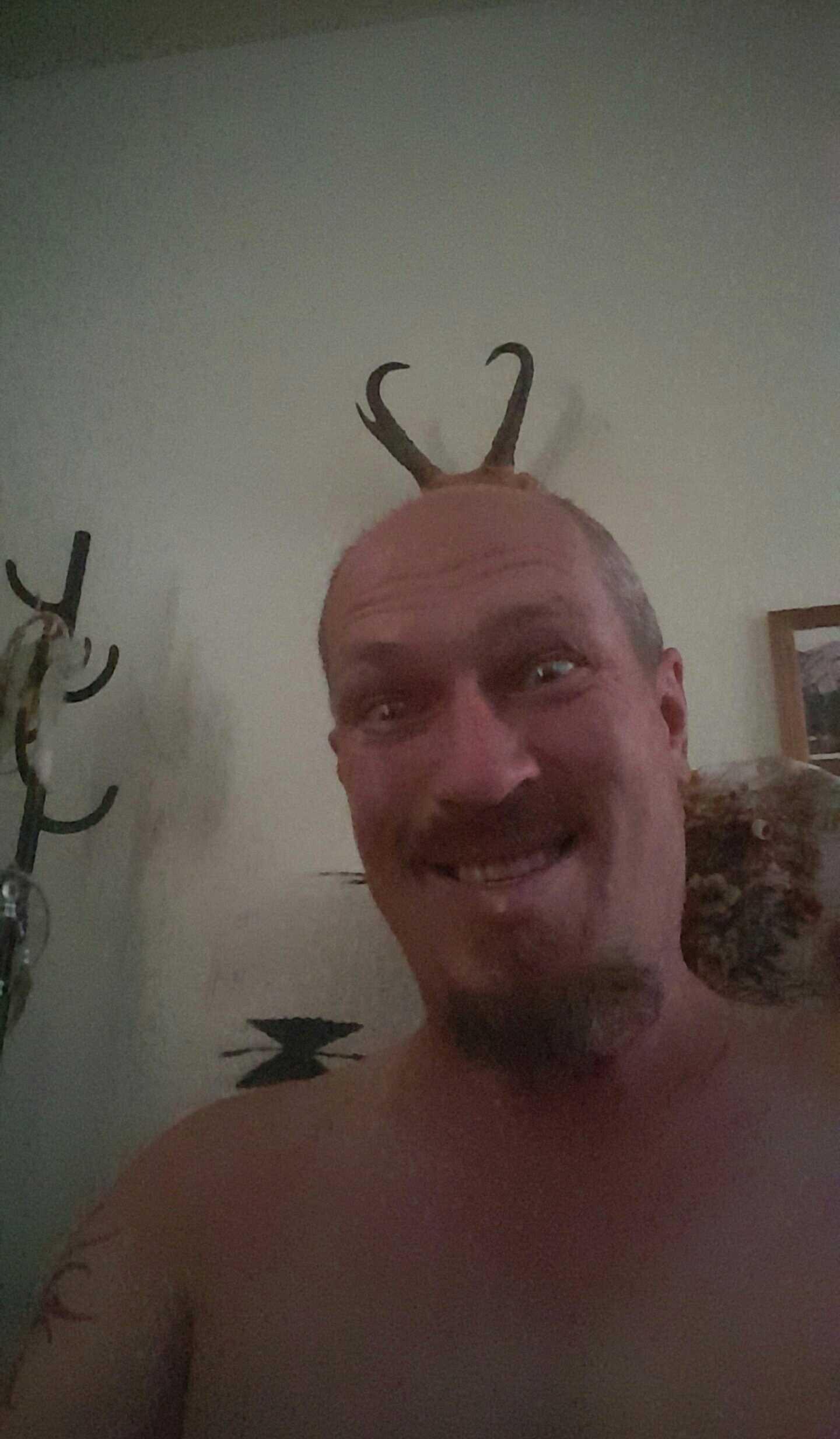 Giant Talon 27.5: A man with a playful expression posing for a selfie, featuring what appear to be decorative horns on his head. He is shirtless, with a goatee, and is sitting in front of a light-colored wall. There are hooks in the background, suggesting a casual indoor setting.