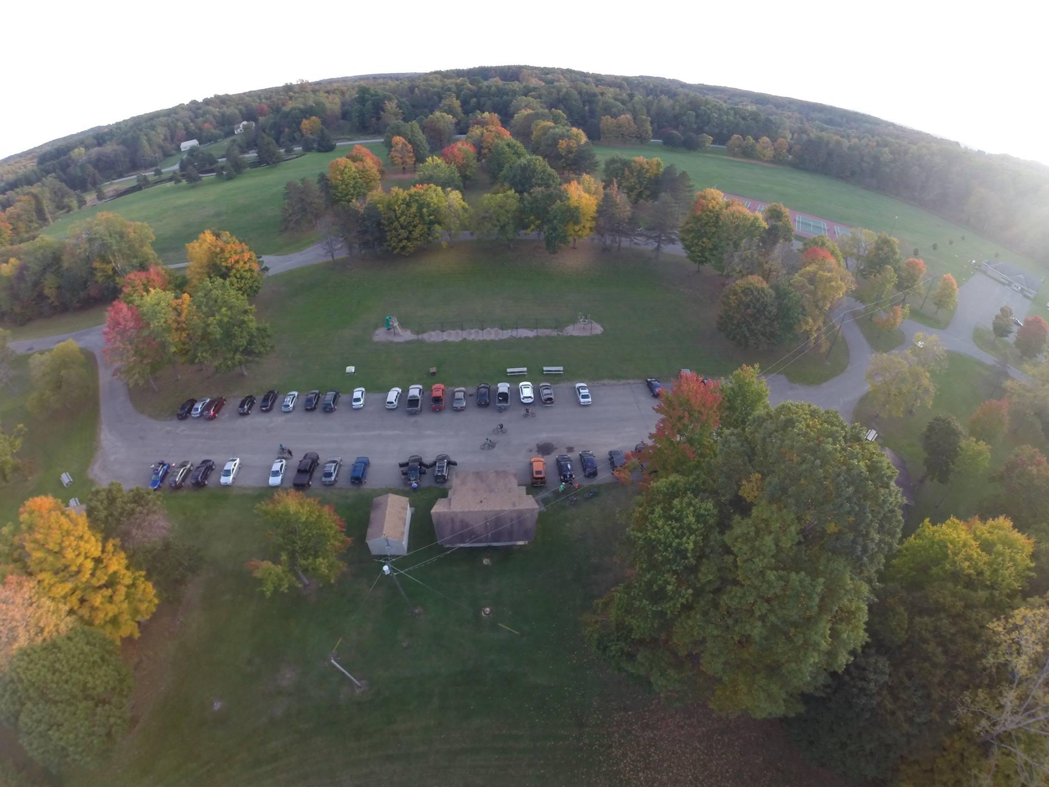 Aerial view of a park during autumn, featuring a parking lot filled with various vehicles, surrounded by trees with colorful fall foliage. Open grassy areas and walking paths are visible, along with a small building and playground equipment in the background. The Full Sprague Experience mountain bike trail.