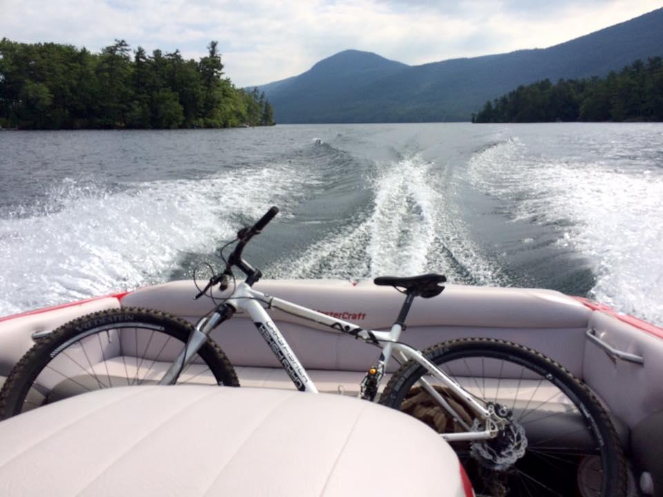 Gary Fisher Paragon: A white mountain bike resting on the edge of a boat with a scenic view of a lake, mountains, and tree-lined shores in the background. The boat is cruising on the water, leaving a wake behind. The sky is partly cloudy.