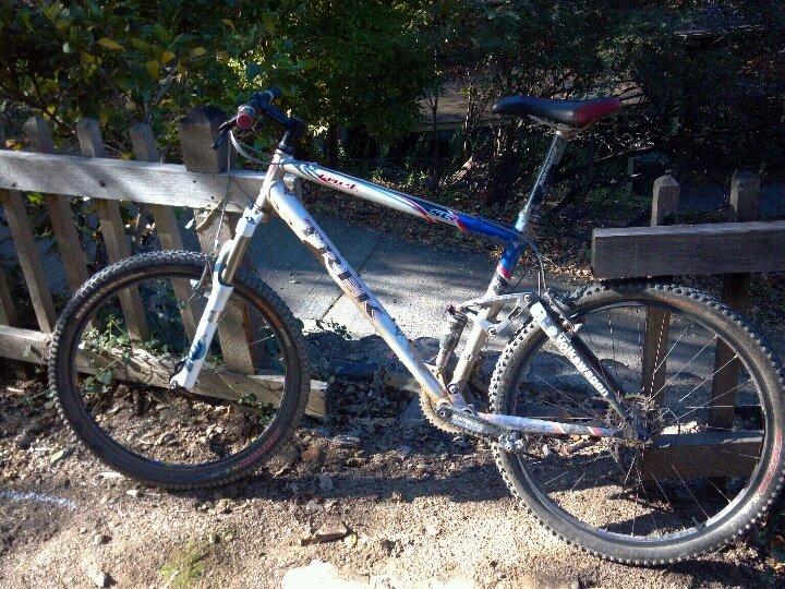Trek Fuel EX: A mountain bike leaning against a wooden fence, with a gravel path and lush greenery in the background. The bike features a predominantly white frame with colorful accents, thick tires, and a basic suspension system, suggesting it's designed for off-road riding.