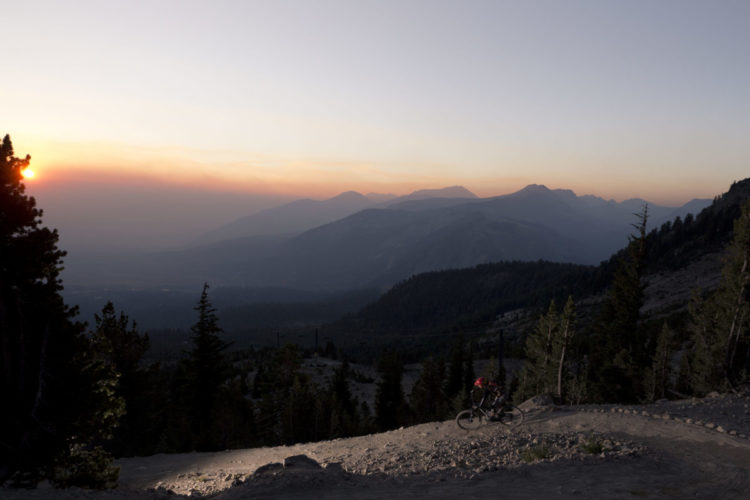 A mountain biker rides along a rocky trail at sunset, surrounded by a scenic landscape of mountains and trees. The sky is painted in hues of orange and pink, creating a beautiful backdrop for the biking adventure.