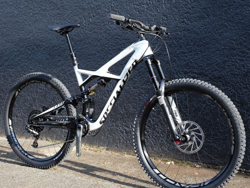 Specialized Enduro Expert Carbon 29: A modern mountain bike with a white and black frame, featuring wide tires and front suspension. The bike is positioned against a textured black wall, showcasing its sleek design and components.