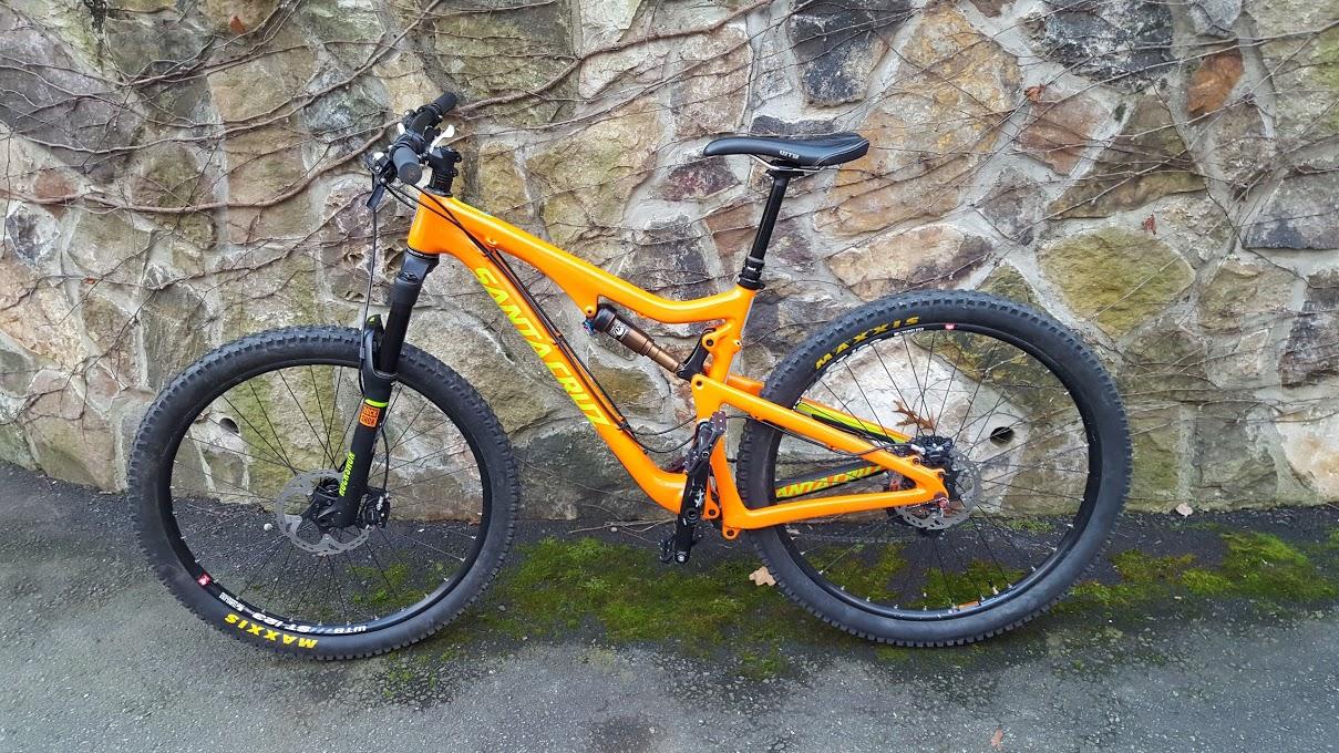 Santa Cruz 5010: A bright orange mountain bike leaning against a textured stone wall, featuring large tires and a black front fork. The bike's frame displays the brand name prominently, and there is some greenery visible at the base.