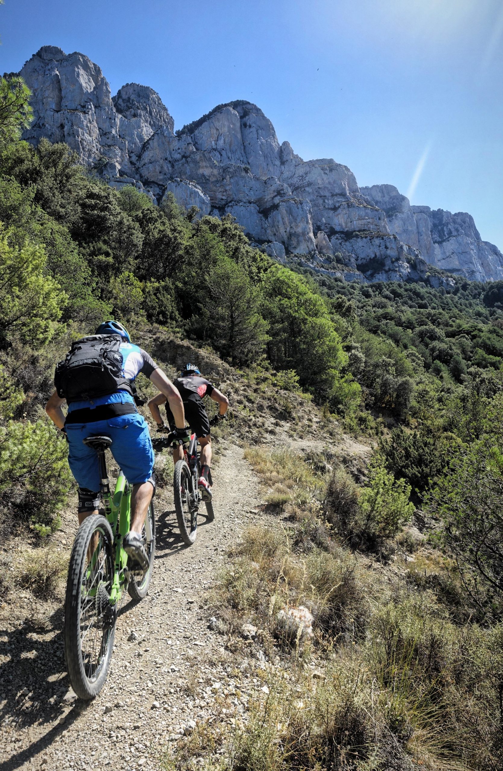 Two mountain bikers ride along a dirt trail surrounded by lush greenery and steep rocky cliffs under a clear blue sky. Maxi Avalanche Trail mountain bike trail.