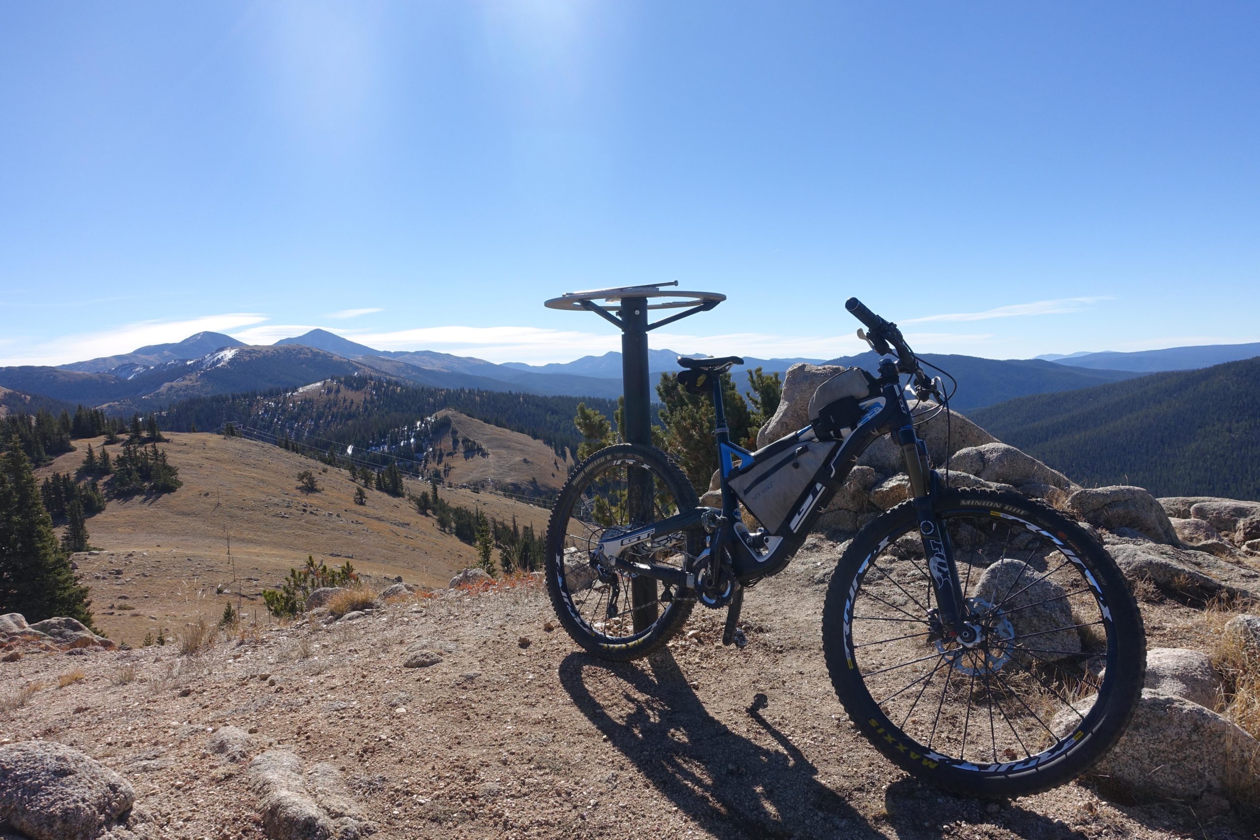A mountain bike parked on rocky terrain with a scenic mountain view in the background. The landscape features rolling hills and pine trees under a clear blue sky. CDT: Monarch Pass to Boss Lake / Hunt Lake Trail mountain bike trail.