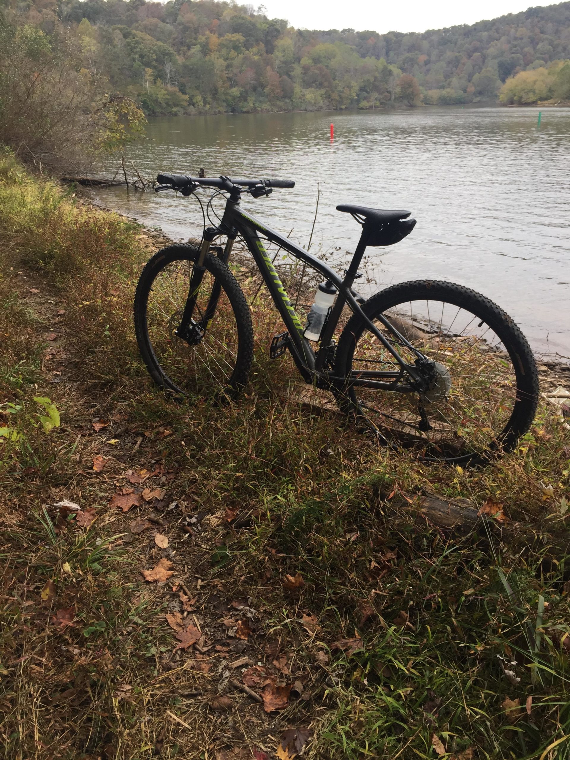 Mountain bike parked on a grassy trail near a calm lake, surrounded by autumn foliage. In the background, green and red markers are visible on the water, indicating navigation points. The scene captures the tranquility of nature in the fall season. Beech Fork State Park mountain bike trail.