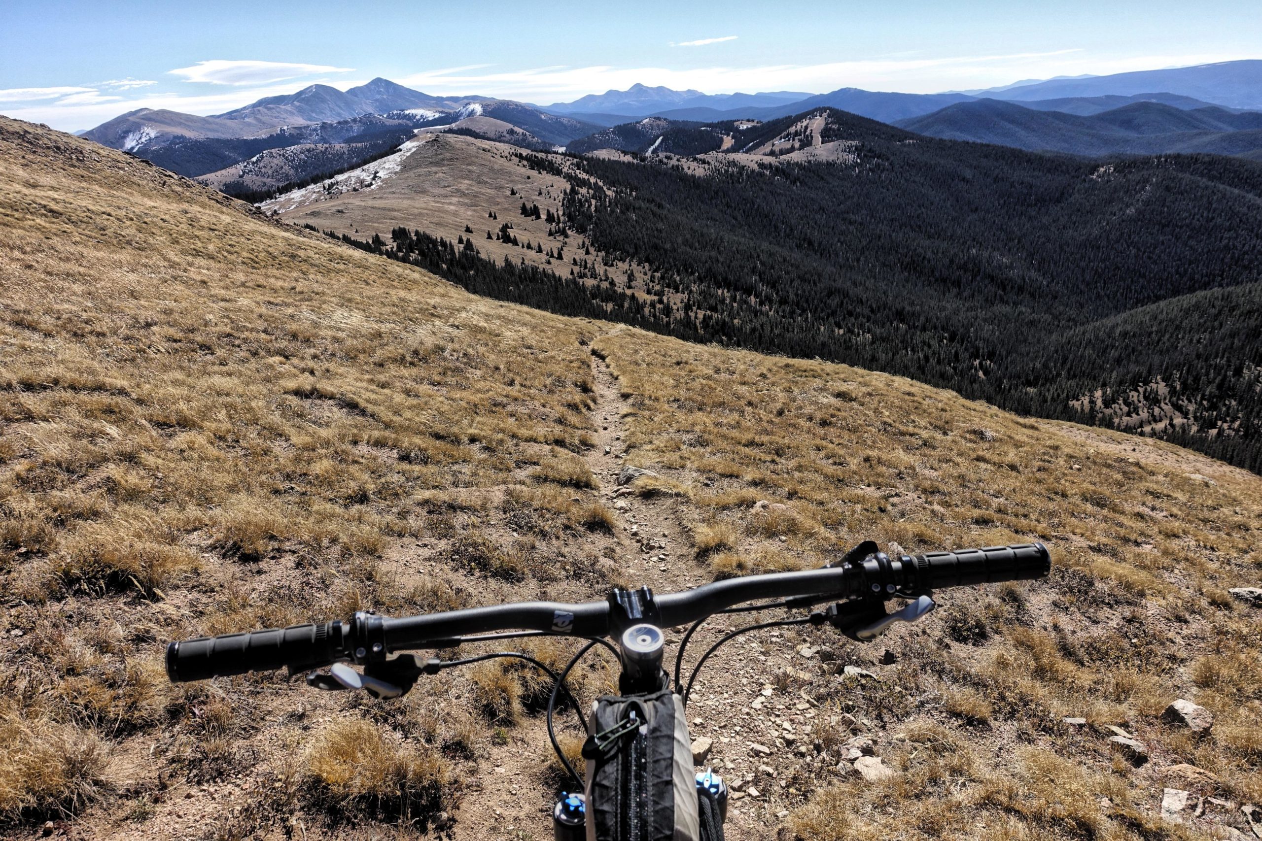 A scenic view from the handlebars of a mountain bike, showing a gravel path winding through golden grasslands and rolling hills, with distant mountains under a clear blue sky. CDT: Monarch Pass to Boss Lake / Hunt Lake Trail mountain bike trail.