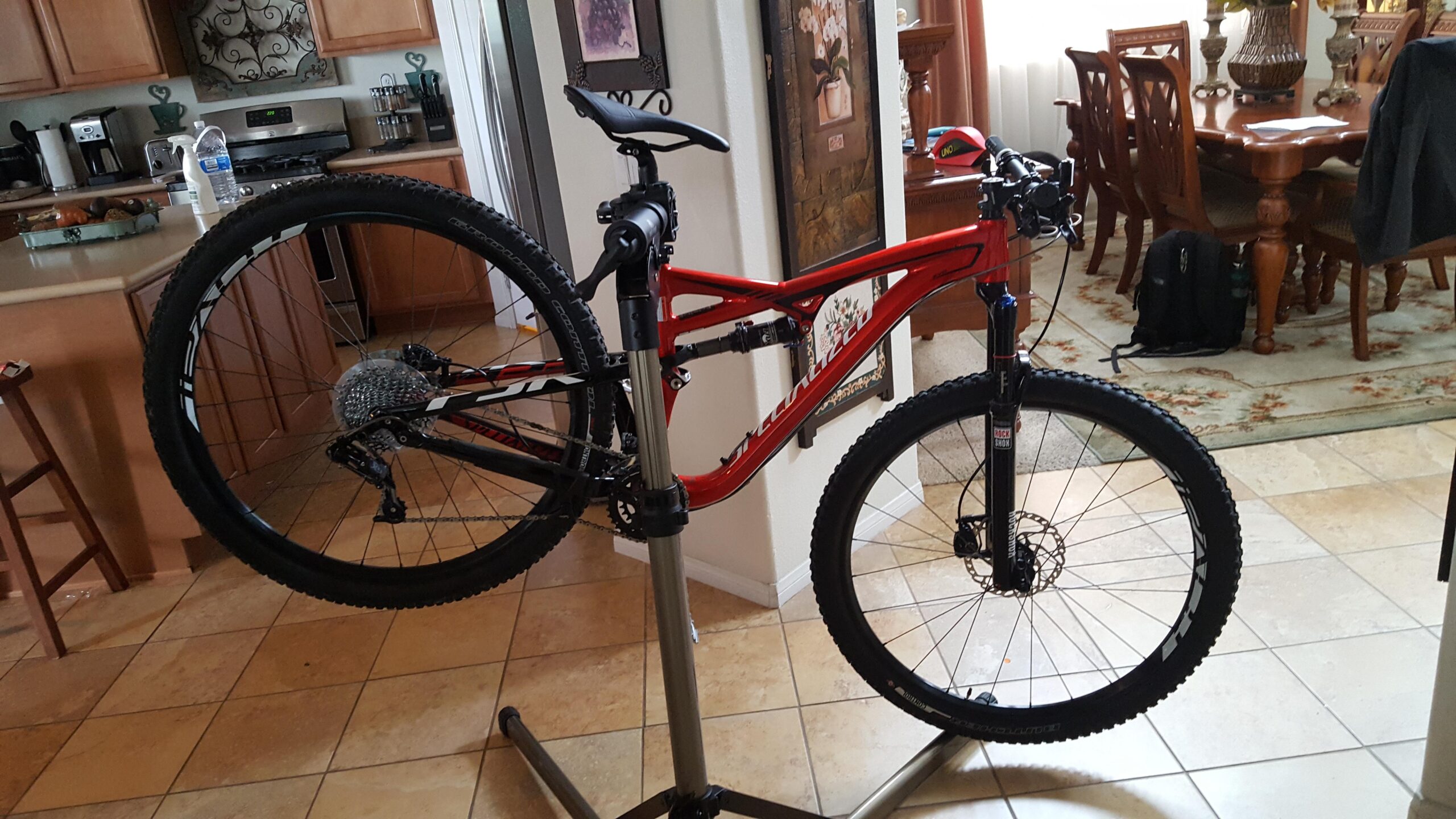 Specialized Camber Comp 29: A red mountain bike is positioned on a bike repair stand in a kitchen, with wooden cabinets and a dining area visible in the background. The bike features thick tires and a visible gear system. A bottle of water and cleaning supplies are on the kitchen counter, and a backpack is placed near the dining table.