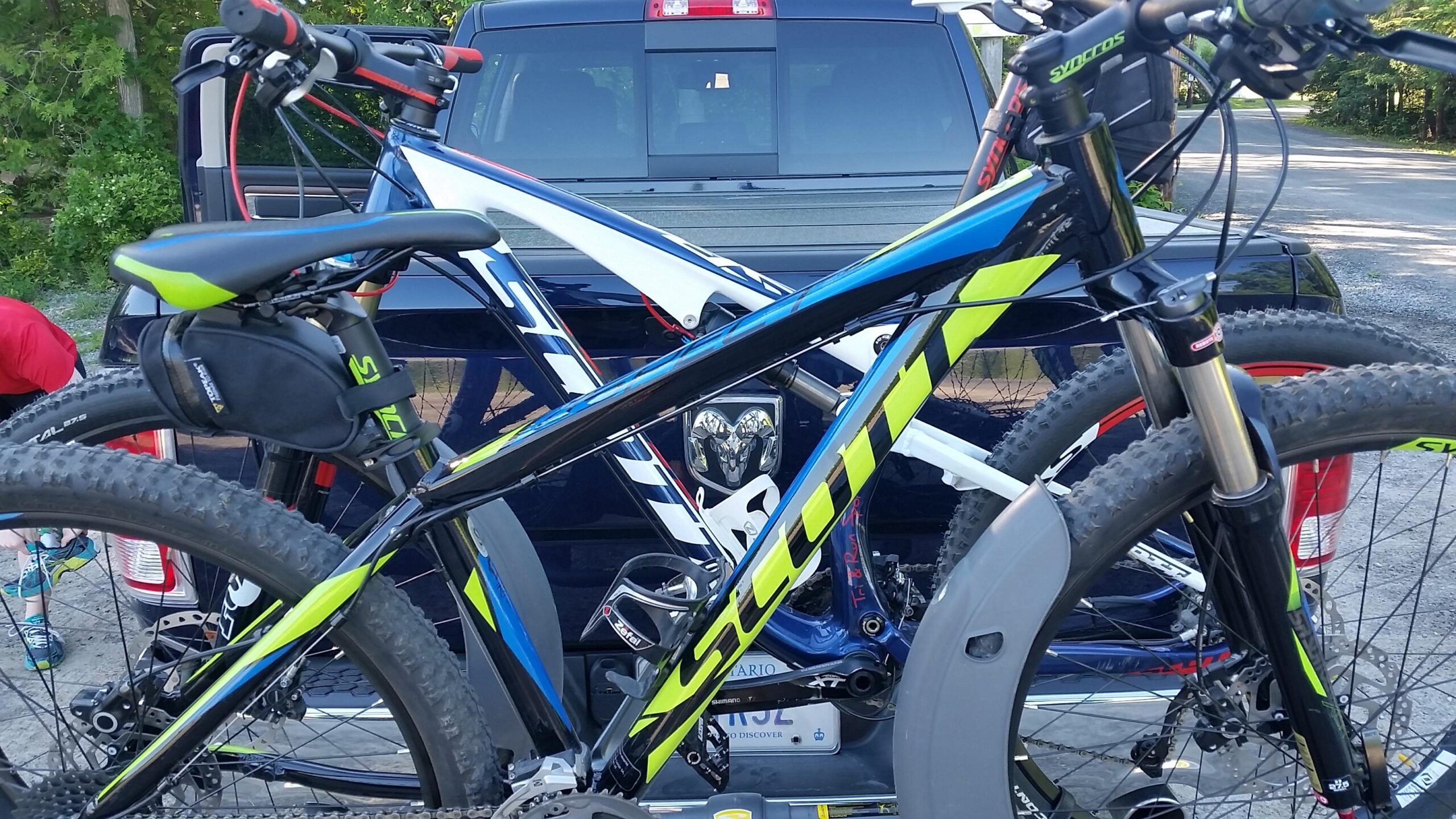 Scott Spark 710: A close-up view of two mountain bikes parked next to a pickup truck. The bikes have distinctive colorful designs, with one prominently featuring black, blue, and lime green colors. The truck's bed and rear are visible in the background, showcasing a logo and part of the tailgate. A person can be seen in the background, adjusting items near the truck. The setting appears to be outdoors, possibly at a trailhead.