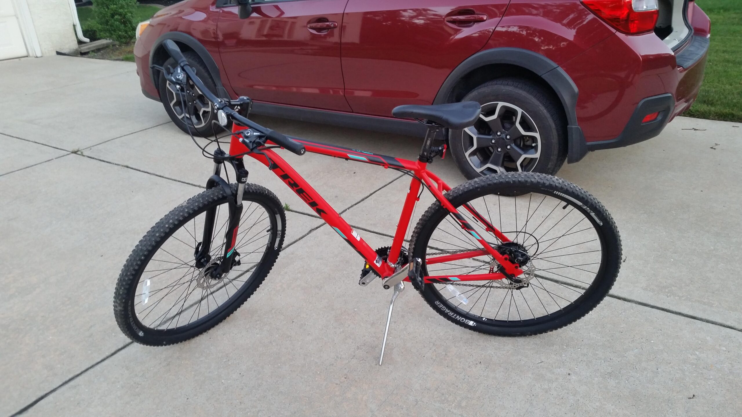 A red Trek mountain bike is parked on a concrete surface, with a red SUV partially visible in the background. The bike features thick tires and a sturdy frame, indicating it is designed for off-road riding.
