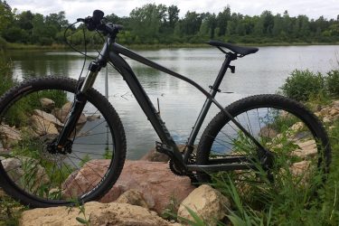 Specialized Rockhopper Comp Disc: A mountain bike resting on large rocks by the edge of a calm lake, surrounded by lush greenery and trees in the background under a partly cloudy sky.